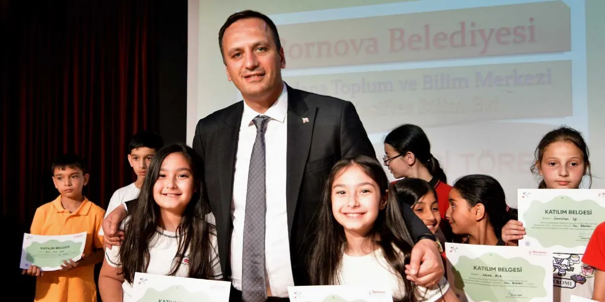 Bornova Belediye Başkanı, bir sahnede düzenlenen törende ellerinde katılım belgeleri bulunan çocuklarla birlikte poz veriyor. Arka planda Bornova Belediyesi Mevlana Toplum ve Bilim Merkezi'ne ait yazıların yer aldığı bir projeksiyon ekranı bulunuyor. Çocuklar ve başkan gülümseyerek kameraya bakıyor.