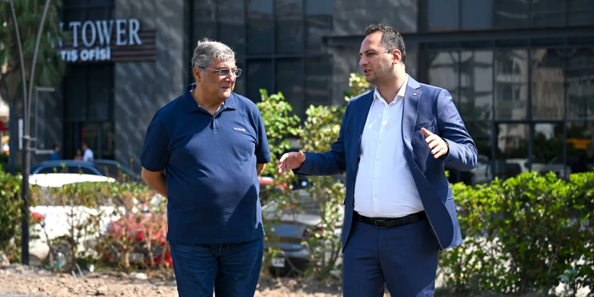 Lacivert polo yaka tişörtlü ve gözlüklü bir erkek ile yanında duran, beyaz gömlek ve lacivert takım elbise giymiş başka bir erkek, dış mekanda bir binanın önünde sohbet ediyor. Takım elbiseli olan kişi ellerini kullanarak bir şeyler anlatırken, yanındaki kişi onu dinliyor. Arka planda ise ağaçlar, park halindeki araçlar ve üzerinde 