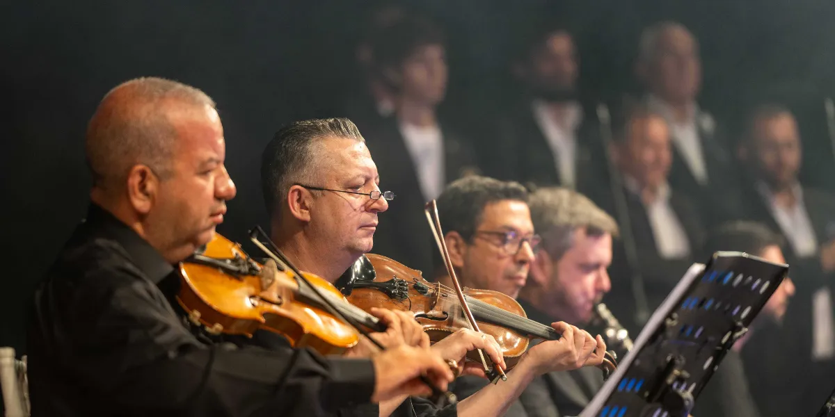 Siyah kıyafetler giymiş bir orkestra grubu, sahne ışıkları altında keman çalarak performans sergiliyor. Ön planda odaklanmış bir şekilde kemanlarını çalan iki müzisyen yer alırken, arka planda ise bulanık bir şekilde diğer orkestra üyeleri ve koro grubu görülüyor.