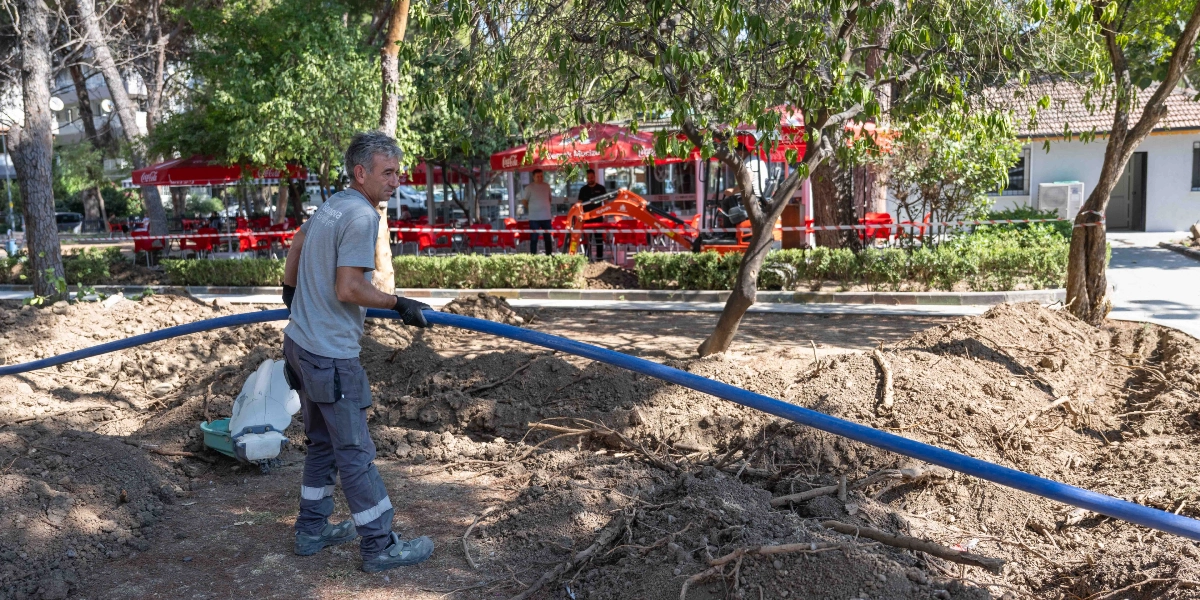 Bornova Belediyesi logolu gri tişört giyen bir işçi, park alanında kazılmış toprağın üzerinde elinde tuttuğu uzun mavi bir sulama borusunu yerleştiriyor. Arka planda ağaçlar, kırmızı şemsiyeli bir kafe alanı ve çalışma alanını çevreleyen güvenlik şeridi görülüyor.