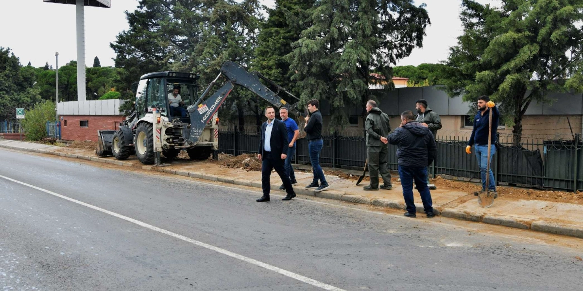 Bornova'da yol kenarındaki kaldırım çalışmasını denetleyen bir grup yetkili ve çalışanlar görülüyor. Siyah takım elbiseli bir kişi önde yürürken, arkasında diğer yetkililer ve ellerinde kürek bulunan işçiler yer alıyor. Arka planda ise sarı renkli bir iş makinesi ve ağaçlıklı bir alan bulunuyor.