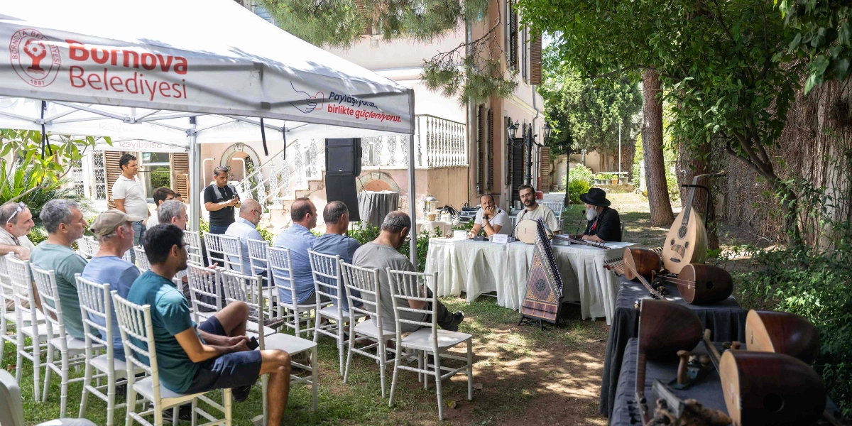 Bornova Belediyesi tarafından düzenlenen bir etkinlikte, ağaçlık bir bahçede beyaz bir çadırın altında toplanan katılımcılar, masa başında oturan konuşmacıları dinliyor. Masada oturan iki kişi ellerinde çalgılarla sunum yaparken, masanın önünde bir kanun ve sağ taraftaki ayrı bir masanın üzerinde ise ud, bağlama ve çeşitli çalgı yapım parçaları sergileniyor.