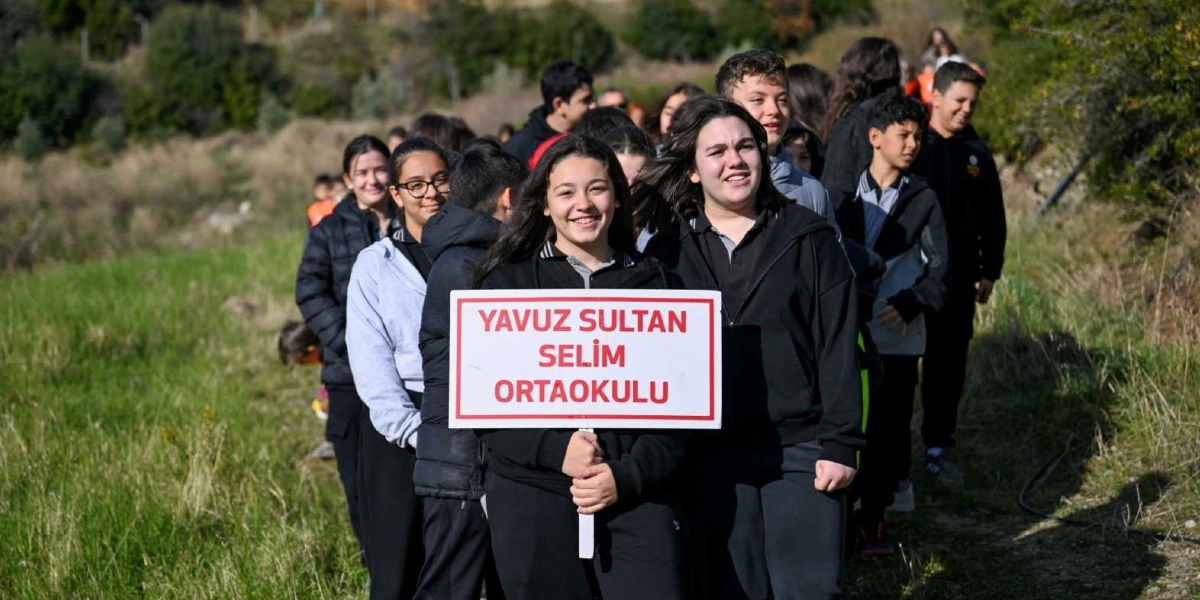 Açık havada, yeşil bir yamaçta yürüyen Yavuz Sultan Selim Ortaokulu öğrencileri, ellerinde okul isimlerinin yazılı olduğu beyaz bir tabela taşıyor. Ön planda gülümseyen iki kız öğrencinin tuttuğu tabelanın arkasında, siyah okul kıyafetleri giymiş diğer öğrenciler tek sıra halinde ilerliyor.