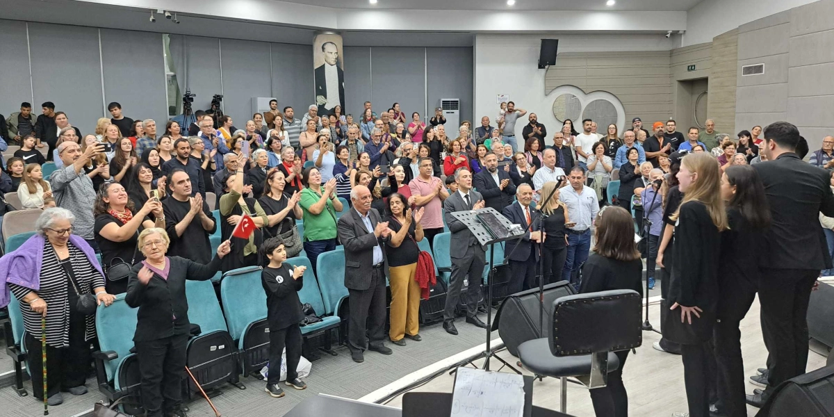 Modern bir konferans salonunda, sahnede şarkı söyleyen bir grup genç ve onları ayakta alkışlayan kalabalık bir izleyici kitlesi yer alıyor. Salonun arka duvarında Mustafa Kemal Atatürk'ün büyük bir portresi asılı. İzleyiciler arasında ellerinde Türk bayrağı tutanlar, cep telefonlarıyla kayıt yapanlar ve sahnedeki performansa eşlik edenler bulunuyor. Salonun koltukları turkuaz renkli olup, ortamda resmi ve duygusal bir atmosfer hakim.