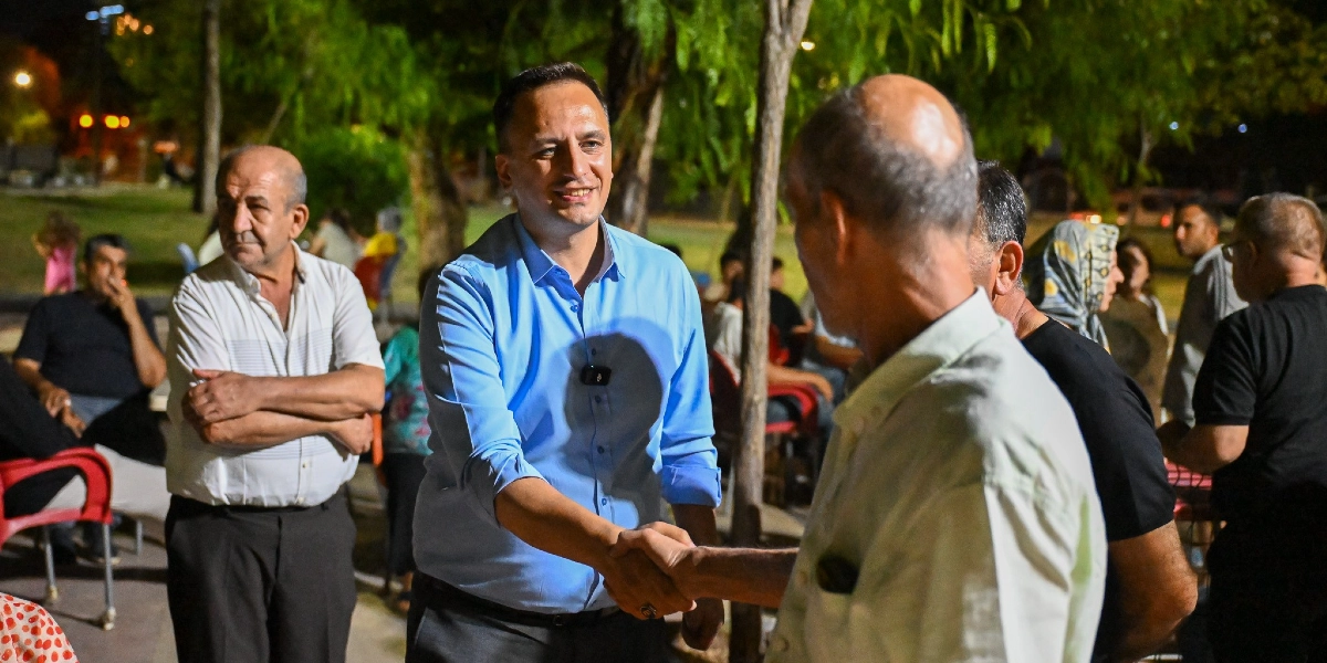 Açık mavi gömlek giymiş bir erkek, akşam saatlerinde dış mekanda bir vatandaşla tokalaşırken gülümseyerek sohbet ediyor. Çevredeki masalarda oturan ve ayakta duran diğer vatandaşlar da bu etkileşimi izliyor.