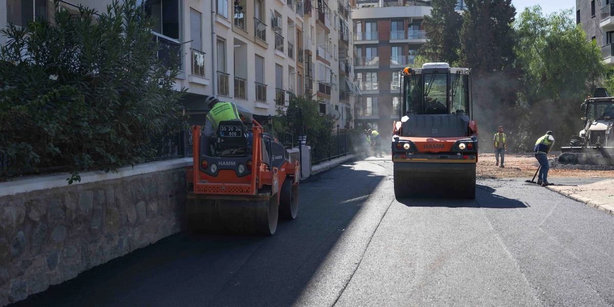 Bornova Belediyesi ekipleri, Erzene mahallesindeki bir sokakta asfalt serme ve yol düzenleme çalışması yapıyor. Çalışma alanında turuncu renkli iki adet Hamm marka silindir iş makinesi, görevli personel ve yol kenarında çok katlı konut binaları yer alıyor.