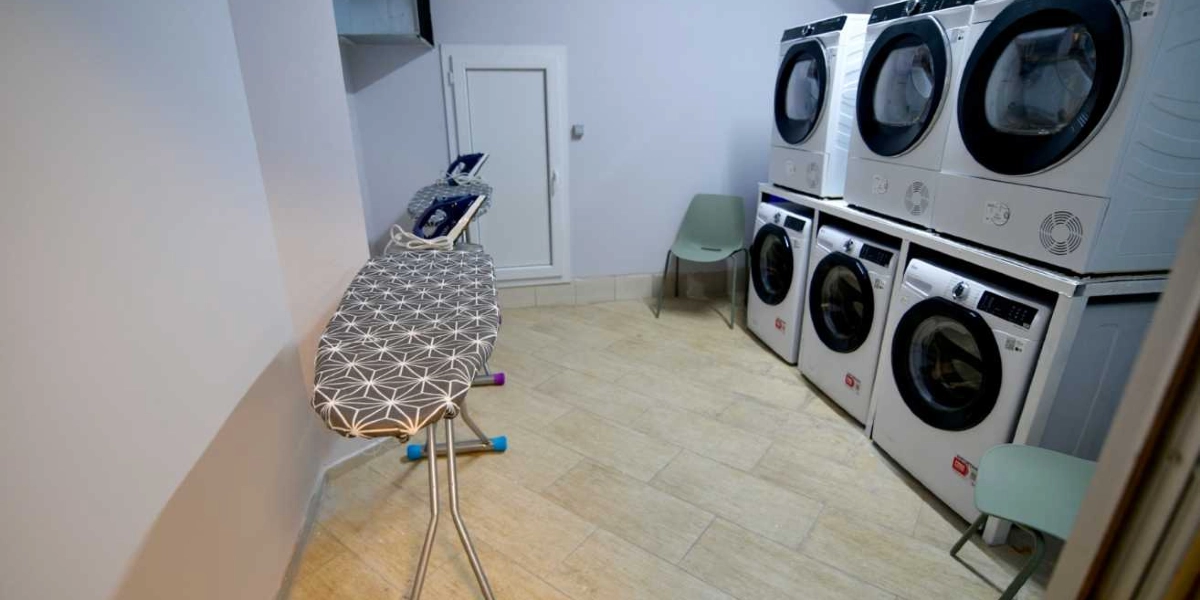 Modern bir çamaşırhanede, üst üste dizilmiş üç kurutma makinesi ve yanlarında üç çamaşır makinesi bulunmaktadır. Odanın sol tarafında geometrik desenli bir ütü masası ve üzerinde ütü yer alırken, köşelerde iki adet açık yeşil plastik sandalye bulunmaktadır. Zemin bej renkli seramiklerle kaplıdır ve duvarlar açık gri tonlarındadır.