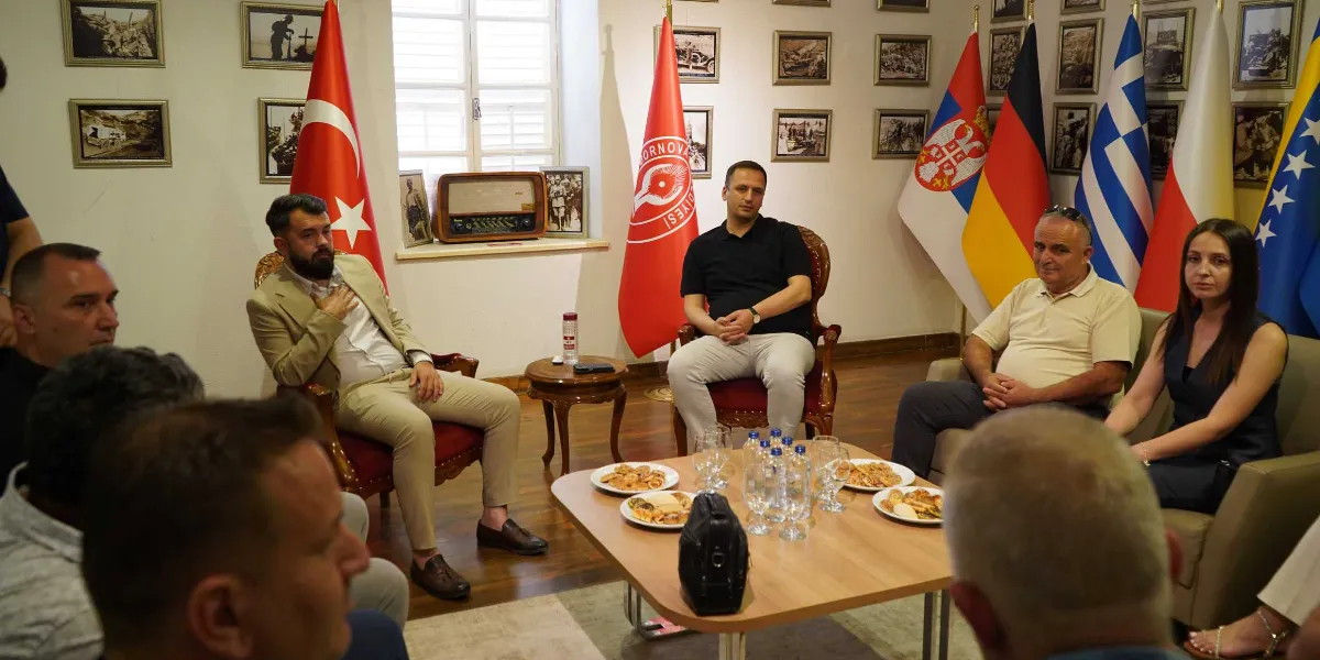 Bornova Belediyesi'nde gerçekleşen toplantıda, karşılıklı oturan bir grup insan yer alıyor. Aralarında Türkiye, Sırbistan, Almanya, Yunanistan, Polonya ve Bosna-Hersek bayraklarının bulunduğu odada, Bornova Belediyesi'nin kırmızı flaması da dikkat çekiyor. Orta kısımdaki masada ikramlıklar ve su bardakları bulunurken, katılımcılar resmi bir görüşme gerçekleştiriyor. Duvarlarda ise tarihi fotoğraflar asılı.