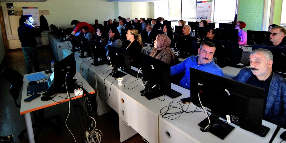 Bir bilgisayar laboratuvarında düzenlenen Bornova Muhtarlar Akademisi eğitimine katılan muhtarlar, masalarda oturmuş bilgisayar ekranlarına bakarak dersi dinliyor. Sınıfın ön tarafında ayakta duran bir eğitmen sunum yaparken, katılımcılar arasında farklı yaş ve cinsiyetlerden muhtarlar yer alıyor. Arka planda üzerinde 