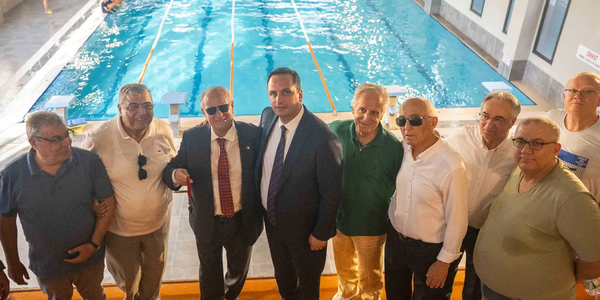 Kapalı bir yüzme havuzunun kenarında yan yana duran ve gülümseyerek kameraya bakan sekiz kişilik erkek grubu. Arka planda kulvarlara ayrılmış masmavi bir yüzme havuzu yer alıyor. Gruptaki kişiler genellikle günlük kıyafetler giymiş olup, ortada yer alan iki kişi takım elbiseli ve kravatlıdır. Sağ tarafta duvarda 