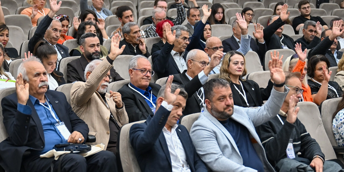 Konferans salonunda oturan çok sayıda katılımcı, bir oylama veya onay sürecini temsilen ellerini havaya kaldırmış durumda. Çoğunluğu resmi kıyafetli olan katılımcıların birçoğunun boynunda etkinlik kartı asılıdır ve salonun genelinde ciddi bir atmosfer hakimdir.