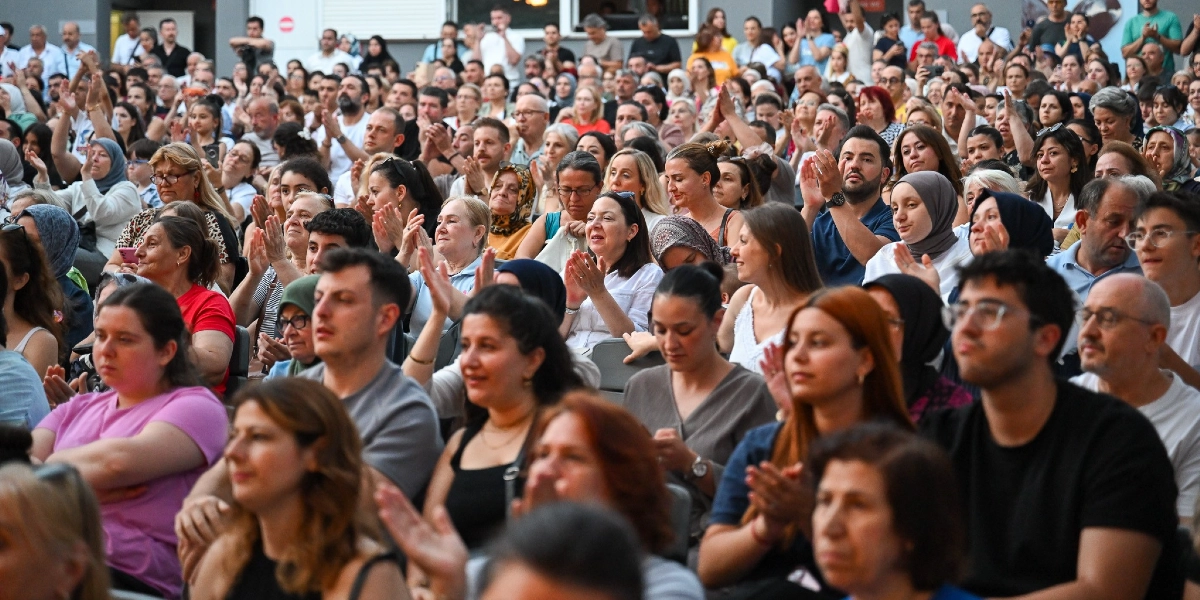 Kapalı bir mekanda, sahnedeki bir gösteriyi izleyen ve alkışlayan çok sayıda kursiyer ve izleyici grubu. Farklı yaş ve tarzlardan oluşan kalabalık, dikkatle sahneye odaklanmış durumda ve birçoğu elleriyle alkış tutuyor.