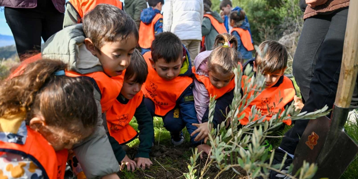 Üzerlerinde turuncu ikaz yelekleri bulunan bir grup anaokulu öğrencisi, açık havada yeni dikilmiş küçük bir fidanın etrafında toplanmış, elleriyle toprağı düzenliyor. Çocukların yanında yetişkinler ve dikim işleminde kullanılan bir kürek yer alıyor.
