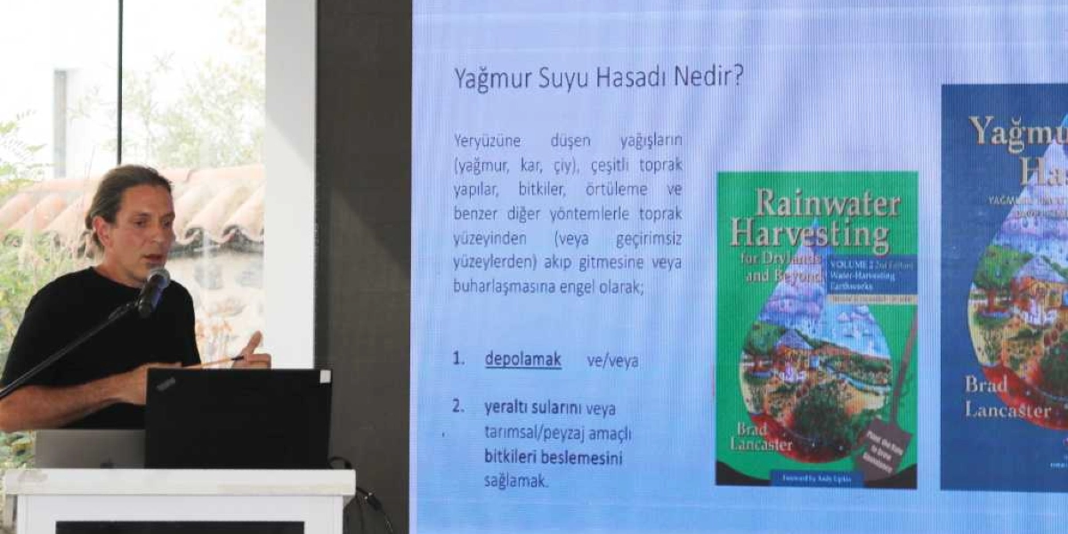 Kürsüde konuşma yapan uzun saçlı bir erkek, arkasındaki dev ekranda 