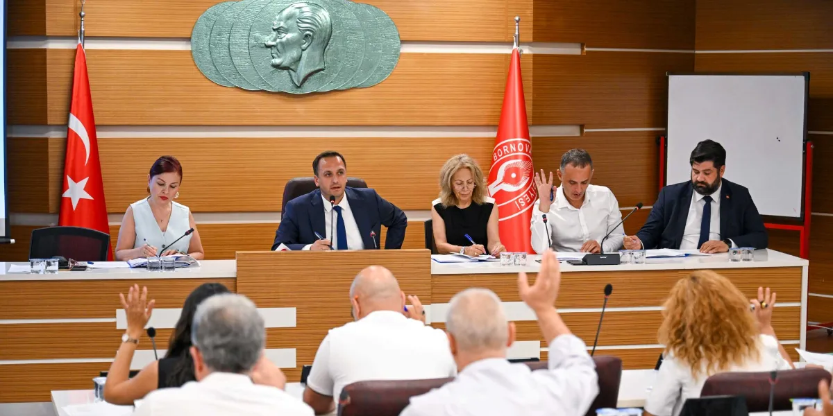 Bornova Belediyesi meclis salonunda, ahşap kaplamalı duvarın önünde yer alan masada oturan beş kişilik bir heyet ve onları dinleyen katılımcılar görülüyor. Duvarın merkezinde Atatürk kabartması, iki yanında ise Türk bayrağı ve Bornova Belediyesi bayrağı yer alıyor. Masadaki heyet üyeleri önlerindeki evraklarla ilgilenirken, salondaki katılımcılardan bazıları ellerini kaldırarak oylamaya veya tartışmaya katılıyor.