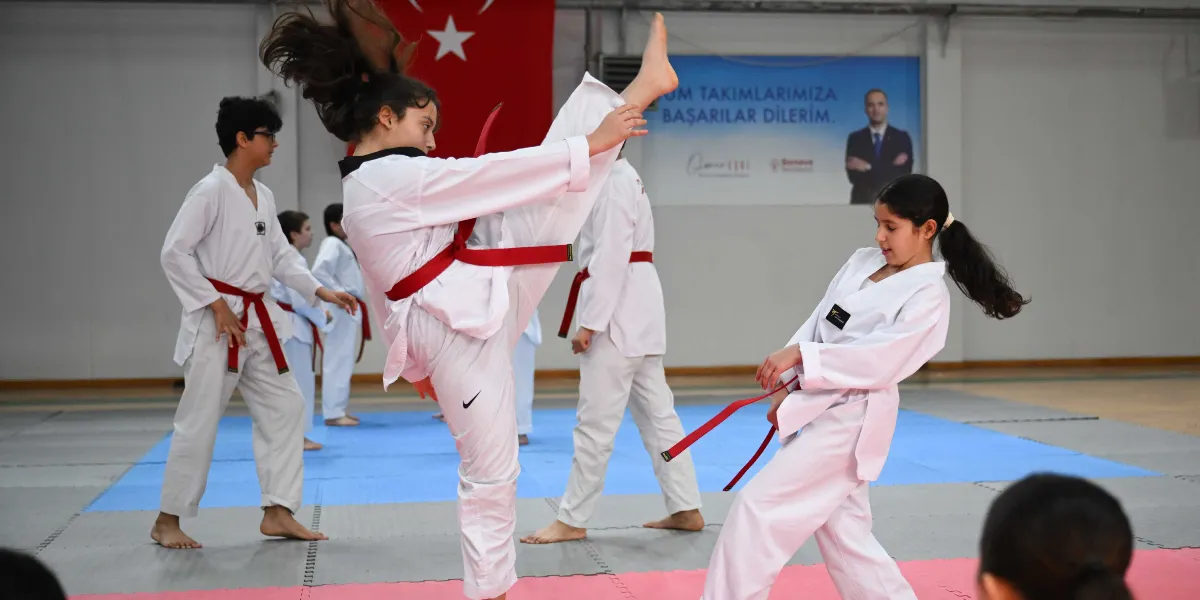 Spor salonunda beyaz tekvando kıyafeti ve kırmızı kuşak giymiş öğrenciler antrenman yapıyor. Ön planda bir kız öğrenci havaya doğru yüksek bir tekme atarken, yanındaki diğer bir kız öğrenci onu izliyor. Arka planda ise diğer öğrenciler benzer kıyafetlerle çalışmaya devam ediyor. Duvarın üzerinde Türk bayrağı ve 