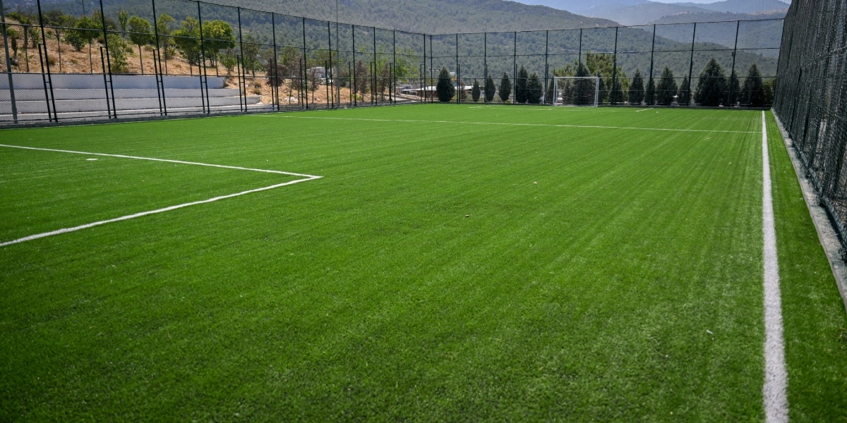 Etrafı yüksek tel örgülerle çevrili, suni çim kaplı boş bir futbol sahası. Sahada beyaz çizgiler belirgin şekilde görülürken, arka planda ağaçlar ve dağ manzarası yer alıyor.