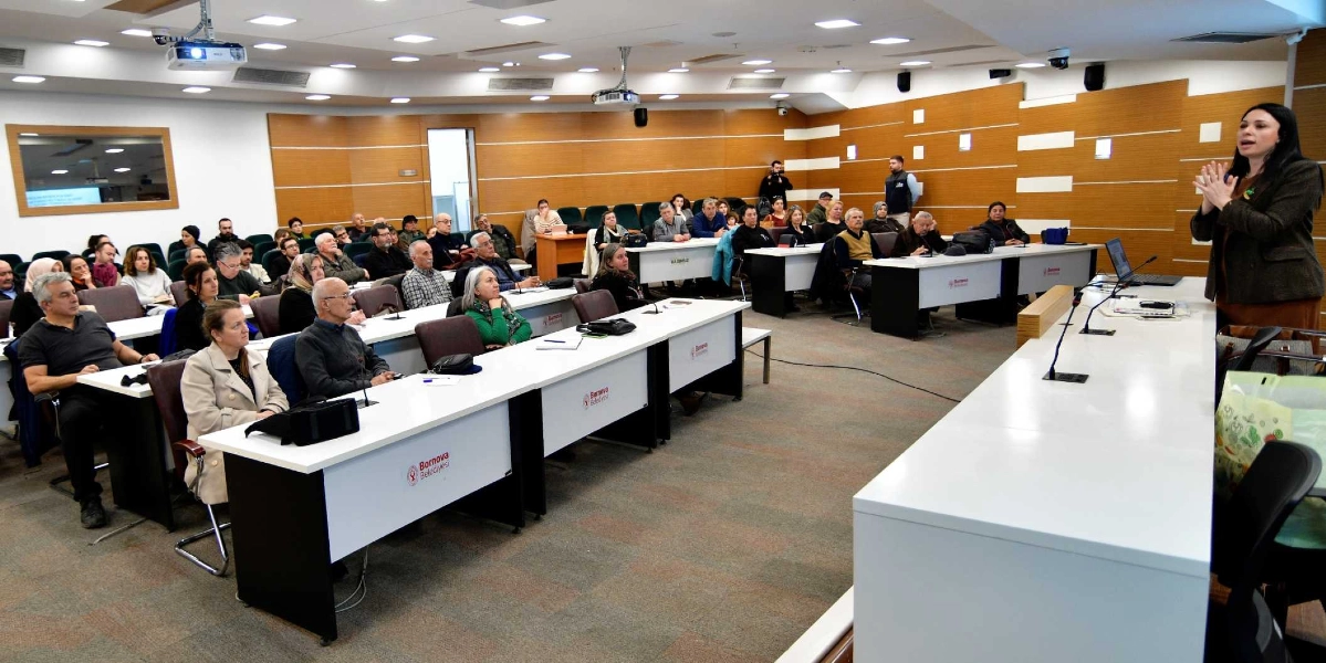 Bornova Belediyesi'ne ait bir konferans salonunda, kürsüde konuşma yapan bir kadın ve onu dinleyen katılımcıların yer aldığı bir toplantı. Katılımcılar, beyaz masaların olduğu sıralarda oturarak sunumu takip ediyor. Salonun duvarları ahşap panellerle kaplı ve tavanda çok sayıda aydınlatma bulunuyor.