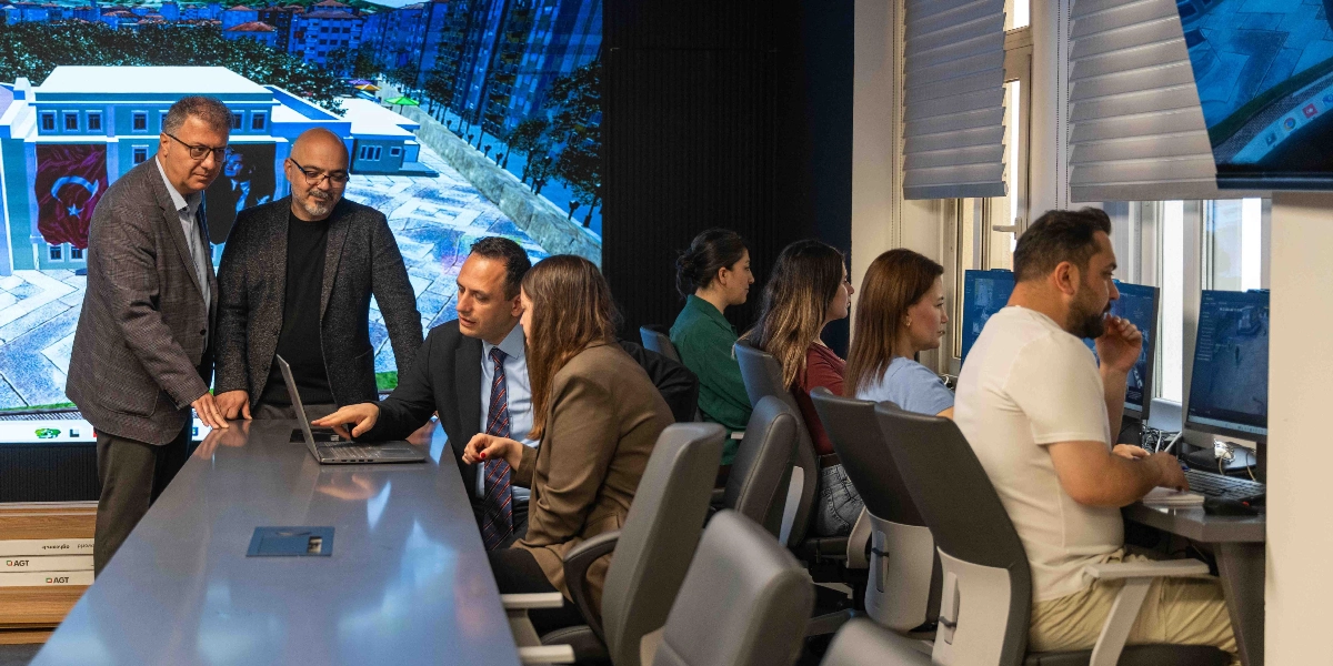 Modern bir ofis ortamında, arka planda büyük bir ekranda dijital bir şehir planlama görseli yansıtılırken, ön planda bir grup çalışan toplantı masası etrafında bir dizüstü bilgisayarı inceleyerek çalışıyor. Masanın başında takım elbiseli üç erkek ve bir kadın iş üzerine konuşurken, masanın devamında ve yan taraftaki bilgisayar istasyonlarında diğer çalışanlar ekranlarını takip ediyor. Ofis, profesyonel bir çalışma atmosferini yansıtıyor.