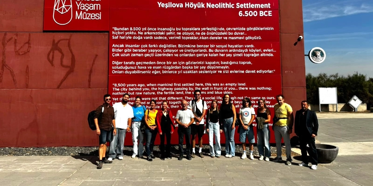 Yeşilova Höyük Neolitik Yerleşimi'ni tanıtan kırmızı renkli büyük bir duvarın önünde yan yana duran, farklı yaş ve giyim tarzlarına sahip 13 kişilik bir grup. Duvarın üzerinde müzenin logosu, 