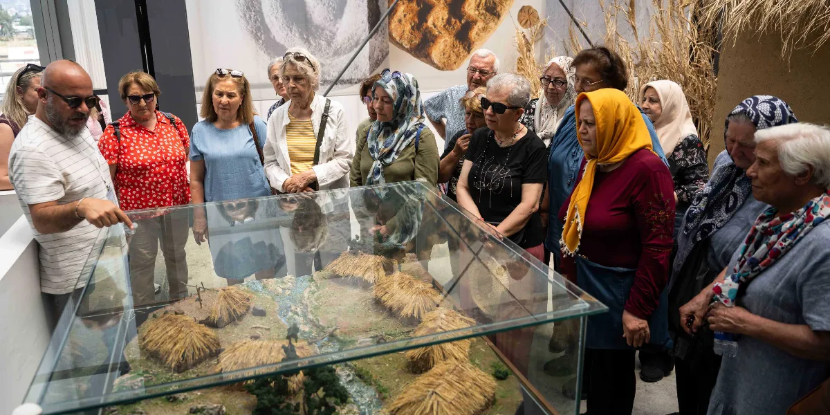 Müzede, sakallı ve gözlüklü bir rehberin, cam bir vitrin içindeki antik köy maketini anlattığı sırada etrafına toplanmış, çoğunluğu kadınlardan oluşan bir grup ziyaretçi. Ziyaretçiler dikkatle maketi inceliyor; arka planda ise üzerinde arkeolojik bulguların görselleri bulunan büyük bir duvar panosu yer alıyor.