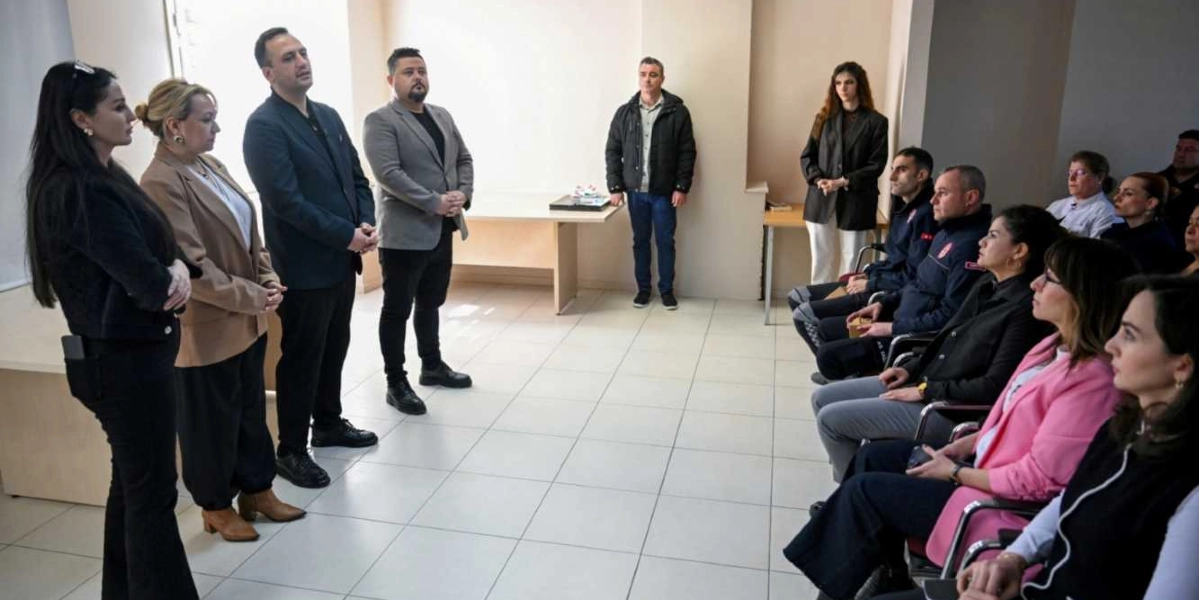 Aydınlık bir odada, bir grup insan toplantı düzeninde bir araya gelmiş durumda. Odanın sol tarafında ayakta duran dört kişilik bir grup, sağ tarafta sandalyelerde oturan ve aralarında üniformalı kişilerin de bulunduğu katılımcılara hitap ediyor. Arka planda ise tek başına ayakta duran bir kişi daha bulunuyor. Katılımcılar dikkatle dinlerken, ortamda resmi bir toplantı havası hakim.