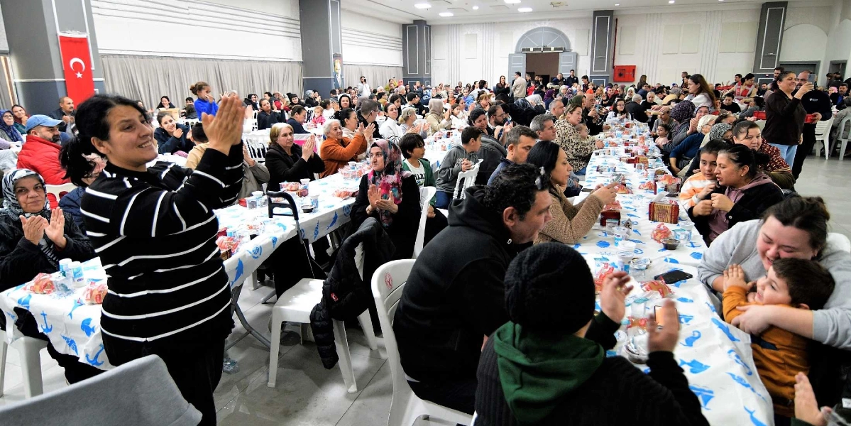 Geniş bir salonda düzenlenen yılbaşı yemeğinde, uzun masalarda oturan kalabalık bir grup Bornovalı vatandaş yer alıyor. Masaların üzeri mavi desenli örtülerle kaplı ve üzerinde yiyecekler bulunuyor. Ön planda, siyah beyaz çizgili bir kazak giymiş bir kadın alkışlarken, sağ tarafta bir kadın kucağındaki küçük bir çocukla ilgileniyor. Salonun duvarında asılı Türk bayrağı dikkat çekerken, katılımcıların birçoğu masalarda oturarak sohbet ediyor ve etkinliğe eşlik ediyor.