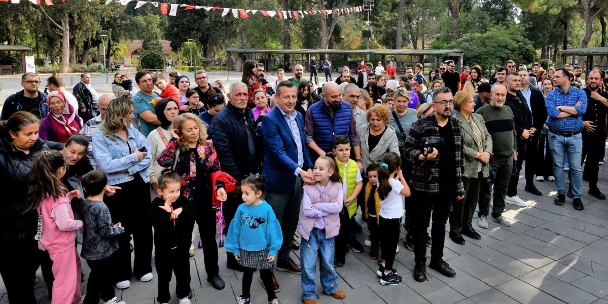 Açık havada, park benzeri bir alanda toplanmış kalabalık bir grup insan, bir etkinliği izlemek üzere yan yana duruyor. Grubun ön planında, mavi ceketli bir adamın yanında duran çocuklar ve yetişkinler dikkat çekiyor. Arka planda ağaçlar ve üzerinde Türk bayraklarının asılı olduğu bir alan görülüyor. İnsanlar genel olarak günlük kıyafetler içinde, meraklı ve neşeli bir ifadeyle bir noktaya doğru bakıyor.