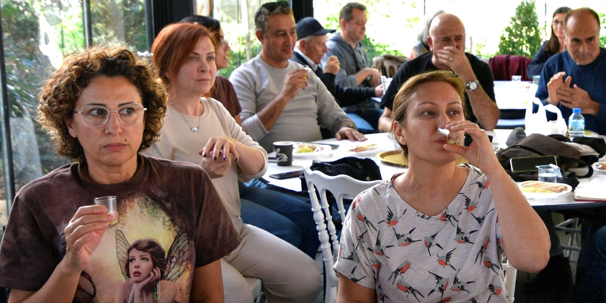 Aydınlık bir iç mekanda, yuvarlak masaların etrafında toplanmış bir grup insan, ellerindeki küçük bardaklarla zeytinyağı tadımı yapıyor. Ön planda, üzerinde peri figürü baskılı kahverengi bir tişört giyen gözlüklü bir kadın ve yanında kuş desenli gri bir tişört giyen, elindeki küçük bardağı ağzına götüren başka bir kadın oturuyor. Arka planda ise çeşitli yaşlardan kadın ve erkekler, masalarda oturarak tadım etkinliğini takip ediyor.