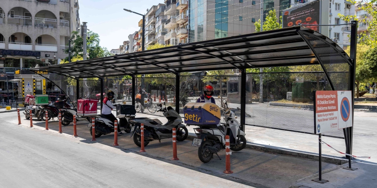 Bornova'da moto kuryeler için güneş ve yağmurdan korunmalarını sağlayan, üstü kapalı ve yanları metal ızgaralı özel bekleme alanı. Alanın içinde park halindeki farklı kurye firmalarına ait motosikletler ve sürücüler bulunuyor. Bekleme alanının girişinde, üzerinde 