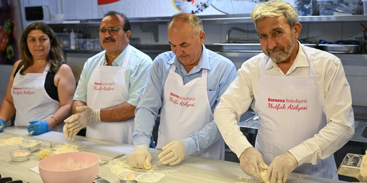 Bornova Belediyesi Mutfak Atölyesi önlükleri giymiş, ellerinde eldivenlerle tezgah başında hamur açan ve makarna hazırlayan dört kişi yan yana duruyor. Endüstriyel bir mutfak ortamında bulunan grup, önlerindeki unlu tezgahta çalışırken kameraya gülümsüyor.