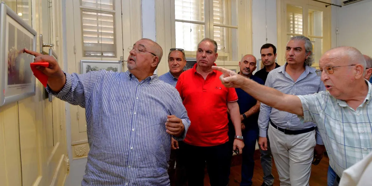 Bir sergi salonunda, duvarda asılı olan Atatürk'e ait tarihi bir fotoğrafı inceleyen ve üzerine konuşan altı kişilik bir grup erkek. Mavi beyaz çizgili gömlekli bir adam, parmağıyla fotoğraftaki bir detayı gösterirken, yanındaki gözlüklü başka bir adam da aynı noktayı işaret ederek açıklama yapıyor. Grubun geri kalanı ise ilgiyle onları dinliyor. Arka planda beyaz ahşap panjurlu pencereler ve duvarda asılı başka fotoğraflar görülüyor.