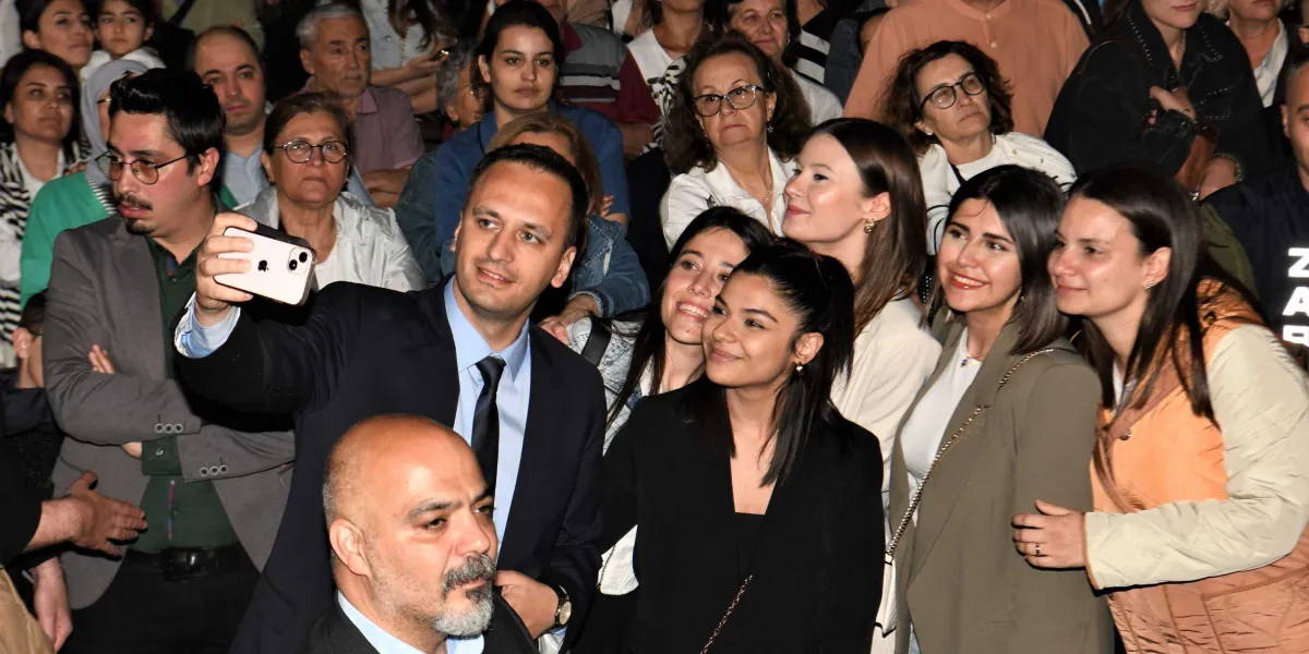 Koyu takım elbiseli bir erkek, kalabalık bir grup insanla birlikte gülümseyerek cep telefonuyla selfie çekiyor. Ön planda yer alan erkek, telefonu tutarken yanında ve arkasında farklı yaş gruplarından kadın ve erkeklerden oluşan bir topluluk bulunuyor. Herkesin kameraya baktığı neşeli bir ortam yansıtılıyor.