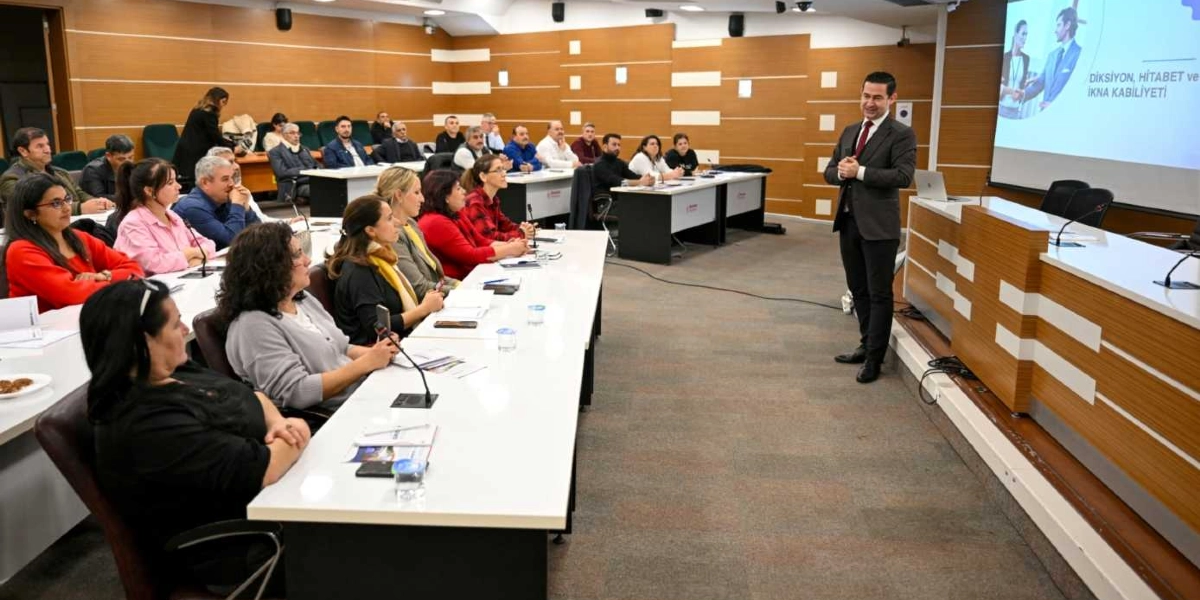 Modern bir konferans salonunda, takım elbiseli bir eğitmen ayakta konuşma yaparken, sıralarda oturan katılımcı grubu onu dinliyor. Salonun önündeki projeksiyon ekranında Diksiyon, Hitabet ve İkna Kabiliyeti yazısı yer alıyor. Katılımcılar masalarda oturmuş, önlerinde not defterleri ve su bardakları ile eğitimi takip ediyor.