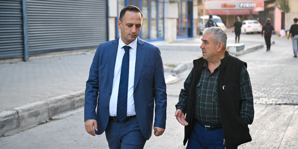 Lacivert takım elbiseli ve kravatlı genç bir adam, yanında ekose gömlek ve siyah yelek giyen orta yaşlı bir adamla birlikte bir sokakta yürürken sohbet ediyor.