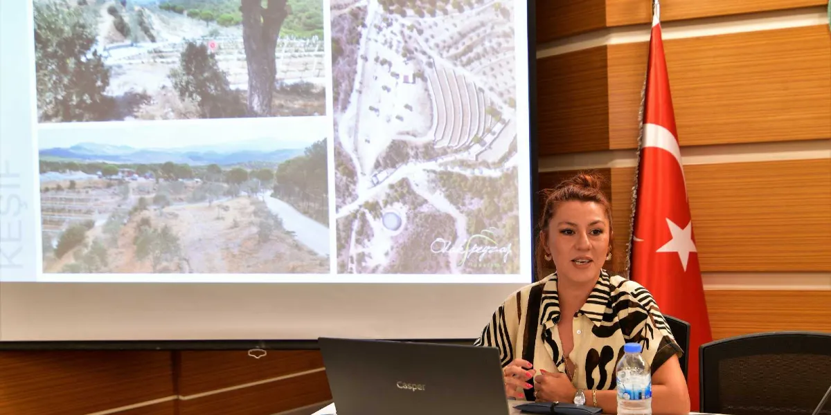 Bir konferans salonunda, arkasındaki projeksiyon perdesinde peyzaj projelerine ait arazi fotoğrafları ve kuş bakışı görünümler yansıyan bir kadın konuşmacı sunum yapıyor. Kadın, üzerinde siyah beyaz desenli bir gömlek ile masada oturuyor ve önünde Casper marka bir dizüstü bilgisayar ile bir şişe su bulunuyor. Salonun sağ tarafında bir Türk bayrağı yer alıyor.
