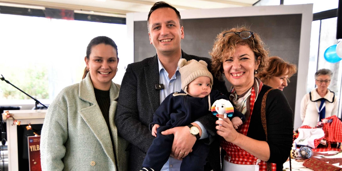 Açık renkli bir kaban giymiş bir kadın, koyu renk ceketli bir adam ve üzerinde kırmızı beyaz ekose bir önlük bulunan kıvırcık saçlı bir kadın yan yana duruyor. Koyu renk ceketli adam, başında bej rengi kulaklıklı bir bere olan küçük bir bebeği kucağında tutuyor. Kıvırcık saçlı kadın elinde küçük, örgü bir oyuncak tutuyor. Arka planda bir stant ve başka insanlar görülüyor.