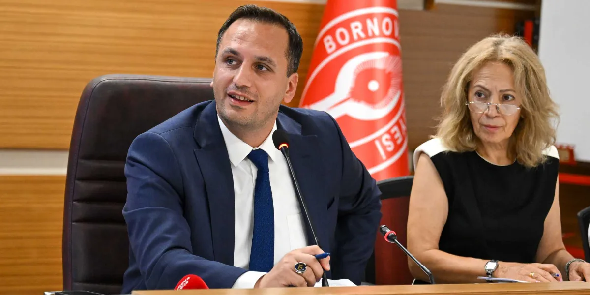 Lacivert takım elbiseli ve mavi kravatlı bir erkek, önündeki mikrofona konuşurken elinde bir kalem tutuyor. Yanında, gözlüklü ve siyah beyaz bir bluz giymiş bir kadın oturuyor. Arka planda Bornova Belediyesi'nin kırmızı beyaz bayrağı yer alıyor.