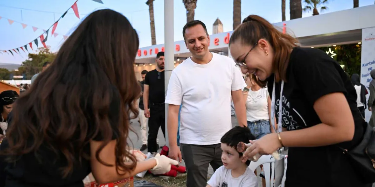 İzmir Enternasyonal Fuarı'ndaki Bornova Belediyesi standında, üzerinde Bornova Belediyesi logosu bulunan siyah tişörtlü görevliler, küçük bir çocuğun yüzüne boya ile desen çiziyor. Etkinliği gülümseyerek izleyen bir baba ve çevredeki diğer ziyaretçilerle birlikte, açık havada kurulan stantta neşeli bir atmosfer yaşanıyor.