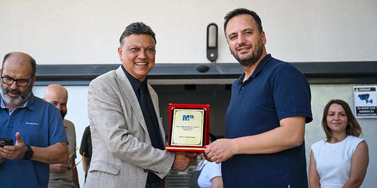 Bej renkli ceket giyen bir kişi, üzerinde MTK logosu ve teşekkür metni bulunan bir plaketi lacivert tişörtlü bir erkeğe takdim ediyor. Arka planda, sol tarafta telefonuna bakan sakallı bir adam ve sağ tarafta gülümseyen bir kadın yer alıyor. Sağdaki cam kapı üzerinde güvenlik kamerası uyarısı içeren bir etiket bulunuyor.