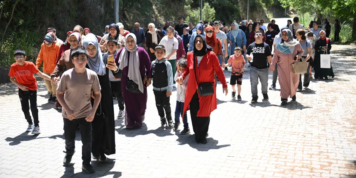 Ağaçlıklı bir yolda, aralarında çocukların, gençlerin ve kadınların bulunduğu kalabalık bir grup, birlikte yürüyüş yapıyor. Grubun ön planında, üzerinde 