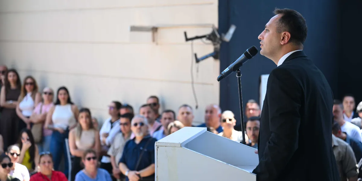 Siyah takım elbiseli bir erkek, açık havada beyaz bir kürsü arkasında durarak mikrofonla konuşma yapıyor. Arka planda ise onu dinleyen kalabalık bir grup insan yer alıyor.
