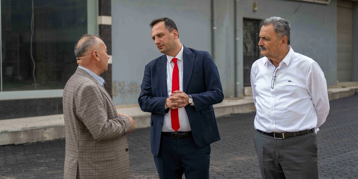 Koyu renkli, yeni asfaltlanmış bir yol üzerinde duran üç erkek, birbirleriyle sohbet ediyor. Ortada lacivert takım elbiseli ve kırmızı kravatlı bir kişi, solunda ekose ceketli bir beyefendi ve sağında beyaz gömlekli, bıyıklı bir beyefendi yer alıyor. Arka planda gri bir bina duvarı görülüyor.