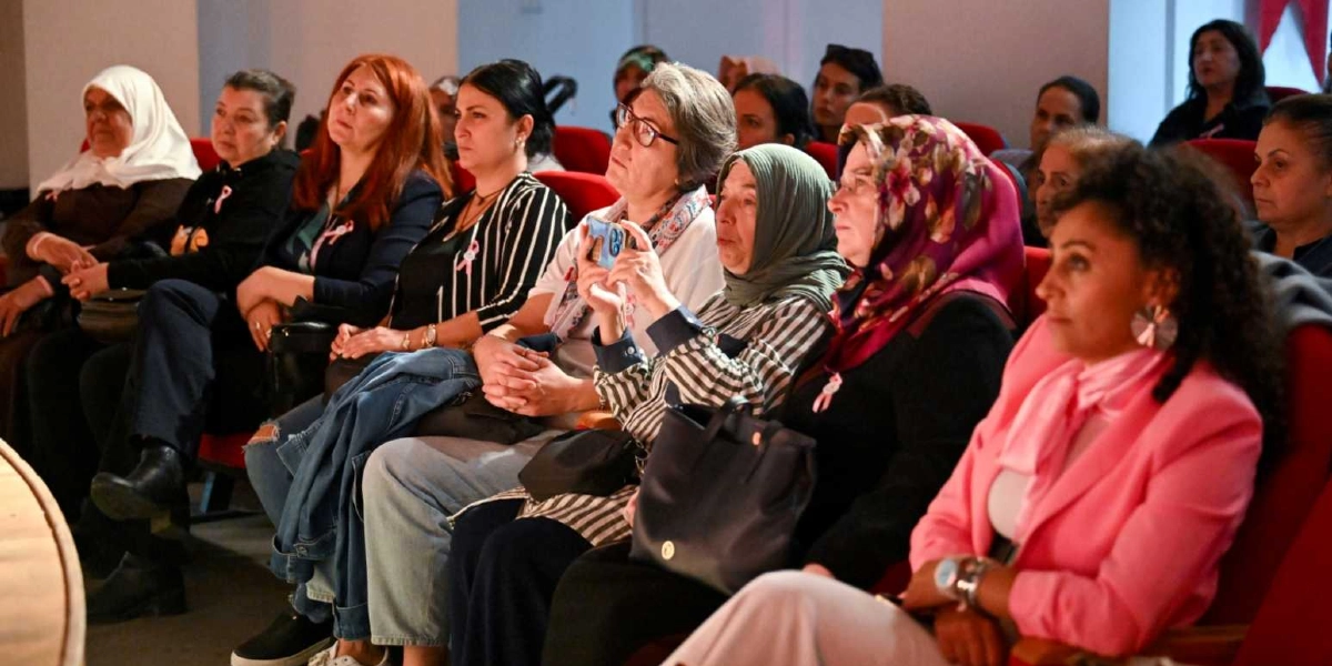 Konferans salonunda kırmızı koltuklarda oturan ve dikkatle sahneyi izleyen bir grup kadın. Birçoğunun yakasında meme kanseri farkındalığını simgeleyen pembe kurdele takılı. Katılımcılardan biri elindeki telefonla sahnenin fotoğrafını çekerken, diğerleri ilgiyle dinliyor. İzleyiciler arasında başörtülü ve başı açık farklı yaş gruplarından kadınlar yer alıyor.