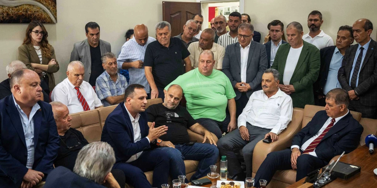 Kapalı bir toplantı odasında, bej renkli koltuklarda oturan ve ayakta duran, çoğunluğu erkeklerden oluşan kalabalık bir grup. Ortada lacivert ceketli bir kişi konuşma yaparken, etrafındaki katılımcılar onu dinliyor. Önlerindeki alçak masada çay bardakları ve atıştırmalıklar bulunuyor. Katılımcıların bir kısmı resmi kıyafetler, bir kısmı ise günlük kıyafetler giymiş durumda.