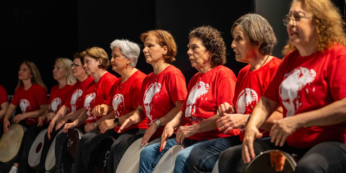 Karanlık bir sahnede yan yana dizilmiş, üzerinde beyaz baskılı kırmızı tişörtler giyen kadınlardan oluşan bir ritim grubu, kucaklarında tuttukları deflerle performans sergiliyor. Orta yaş ve üzeri kadınlar, odaklanmış bir şekilde elleriyle deflere vurarak ritim tutuyor.