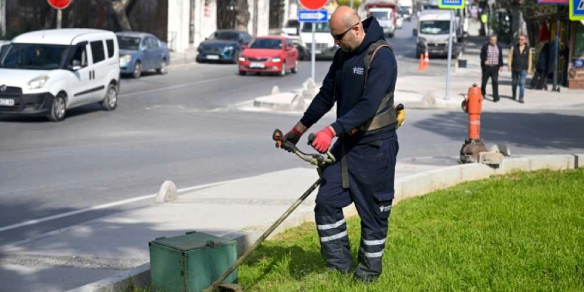 Bornova Belediyesi üniformalı bir işçi, cadde kenarındaki yeşil alanda motorlu tırpan ile çim biçme çalışması yapıyor. Arka planda trafik akışının olduğu bir yol, araçlar ve kaldırımda yürüyen yayalar görülüyor.