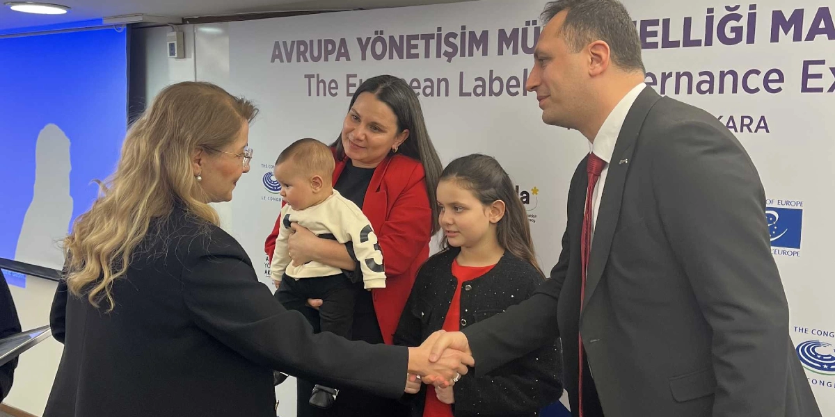 Siyah ceketli bir kadın ile takım elbiseli bir erkeğin tokalaştığı tören sırasında, erkeğin yanında kırmızı ceketli bir kadın, kucağında bir bebek ve yanlarında küçük bir kız çocuğu duruyor. Arka planda Avrupa Konseyi logolarının ve 