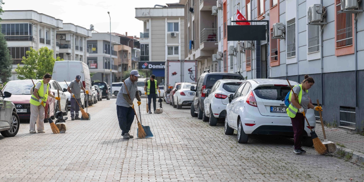 Bornova'da bir sokakta temizlik yapan belediye görevlileri; ellerinde süpürge ve faraşlarla park halindeki araçların çevresini ve kaldırımları temizliyor. Sarı ikaz yelekli çalışanlar, kilit taşı döşeli cadde üzerinde çalışmalarını sürdürüyor.