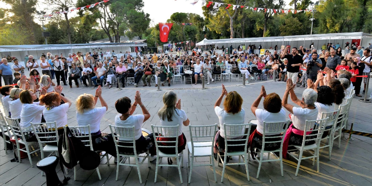 Açık havada düzenlenen bir etkinlikte, beyaz tişörtlü bir grup kadın, sandalyelerde oturarak elleriyle ritim tutuyor. Kadınların önünde, onları izleyen kalabalık bir seyirci grubu ve arka planda beyaz stantlar ile Türk bayrağı yer alıyor. İlber Ortaylı, kalabalığın önünde ayakta durarak konuşma yapıyor.